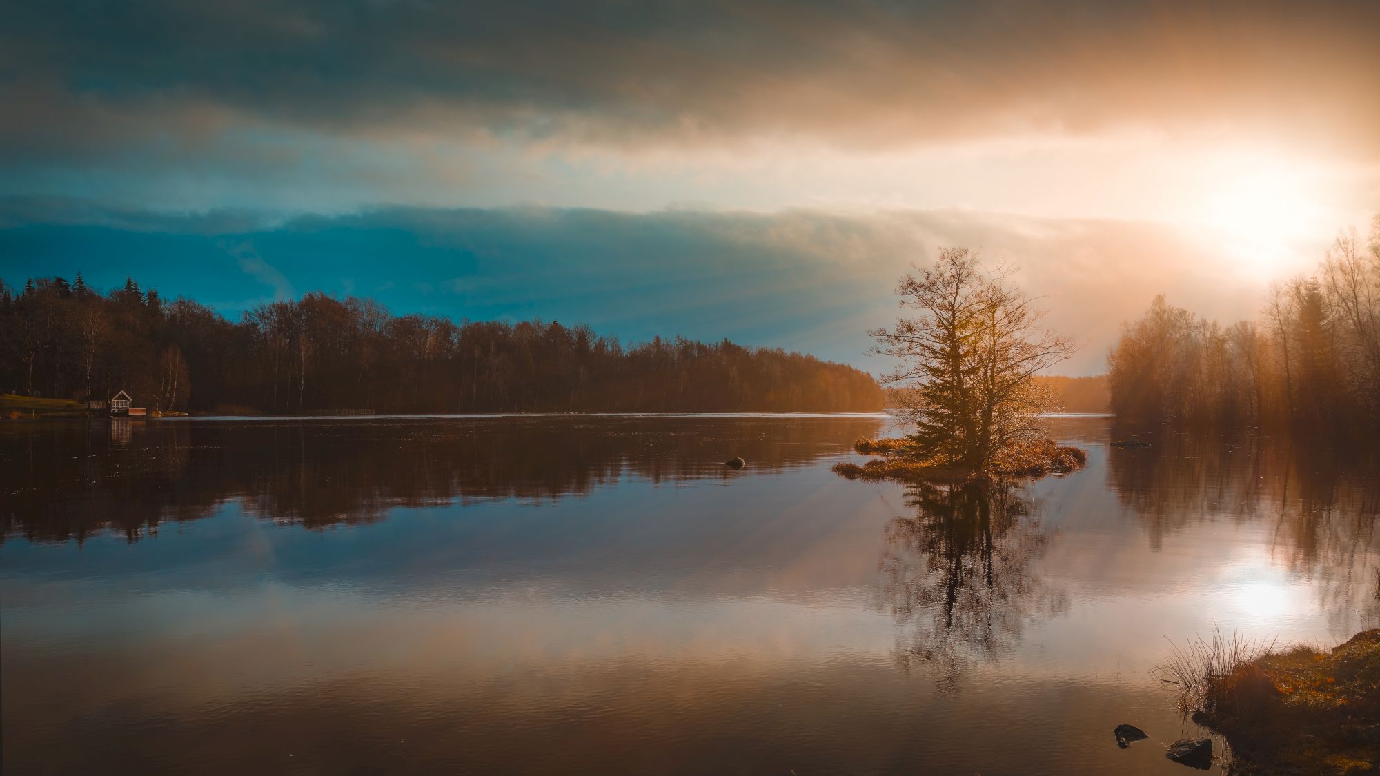 A serene lake scene with a small island, trees, and a vibrant sunset casting warm light on the water, framed by a forest and a cloudy sky.