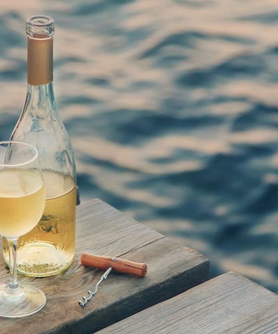 Two filled wine glasses, an opened bottle of white wine, and a corkscrew sit on a wooden dock overlooking gently rippling water, creating a serene ambiance.