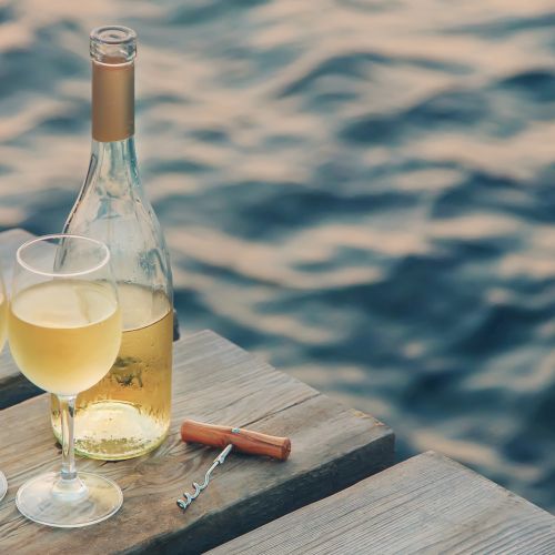 Two filled wine glasses, an opened bottle of white wine, and a corkscrew sit on a wooden dock overlooking gently rippling water, creating a serene ambiance.