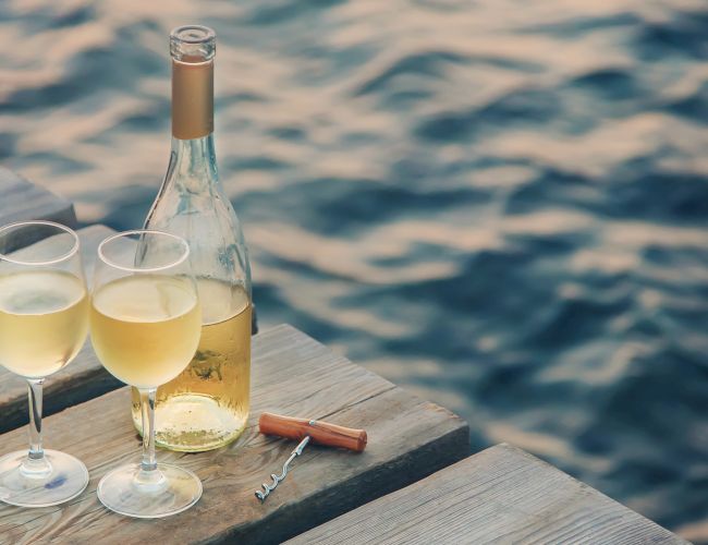 Two filled wine glasses, an opened bottle of white wine, and a corkscrew sit on a wooden dock overlooking gently rippling water, creating a serene ambiance.