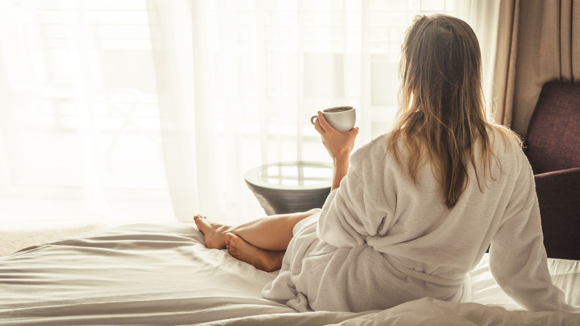 A person in a bathrobe sits on a bed, holding a cup, gazing out a window with sheer curtains.
