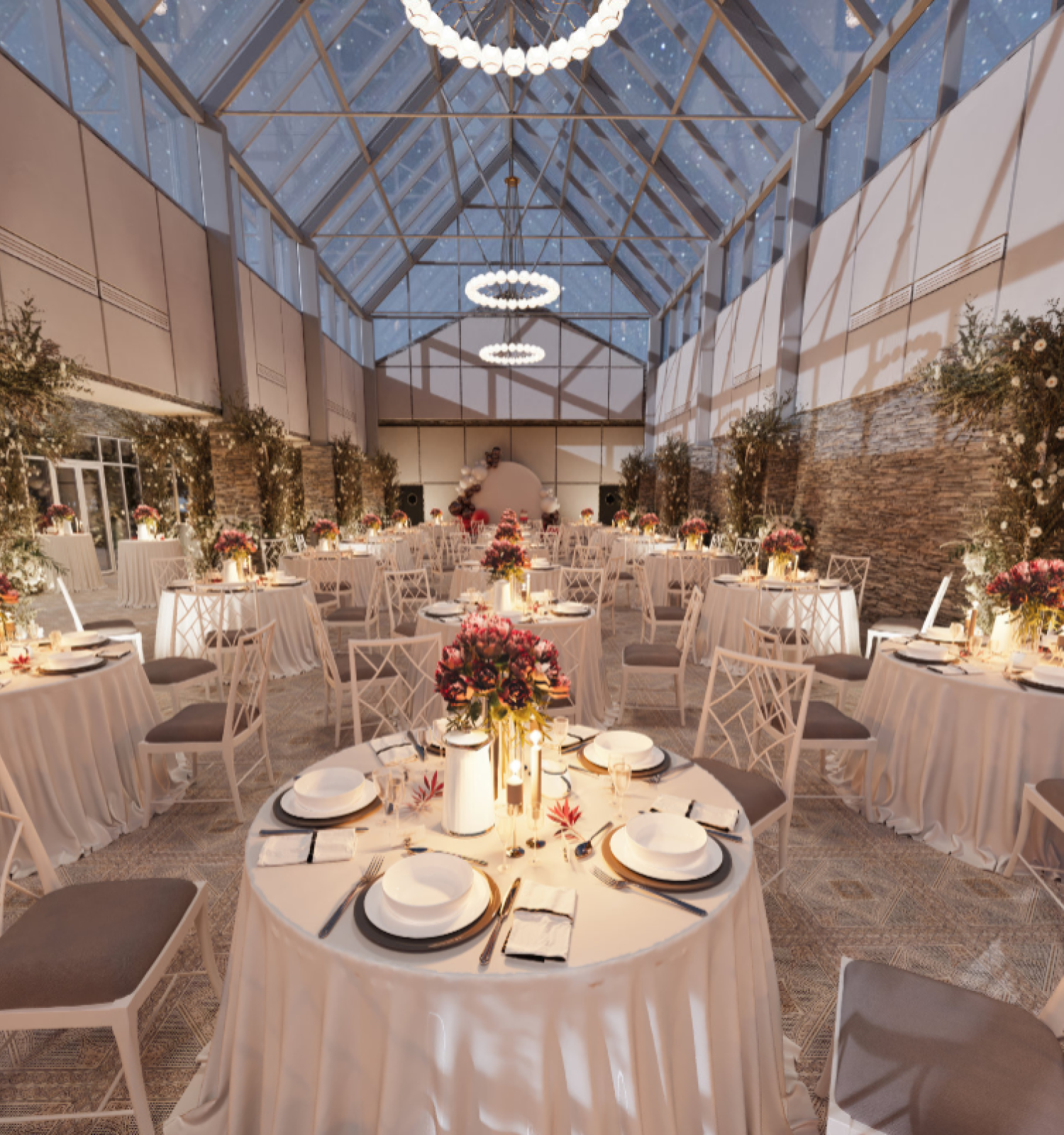 An elegant, indoor event setup with round tables, white tablecloths, floral centerpieces, and chandelier lighting under a glass ceiling.