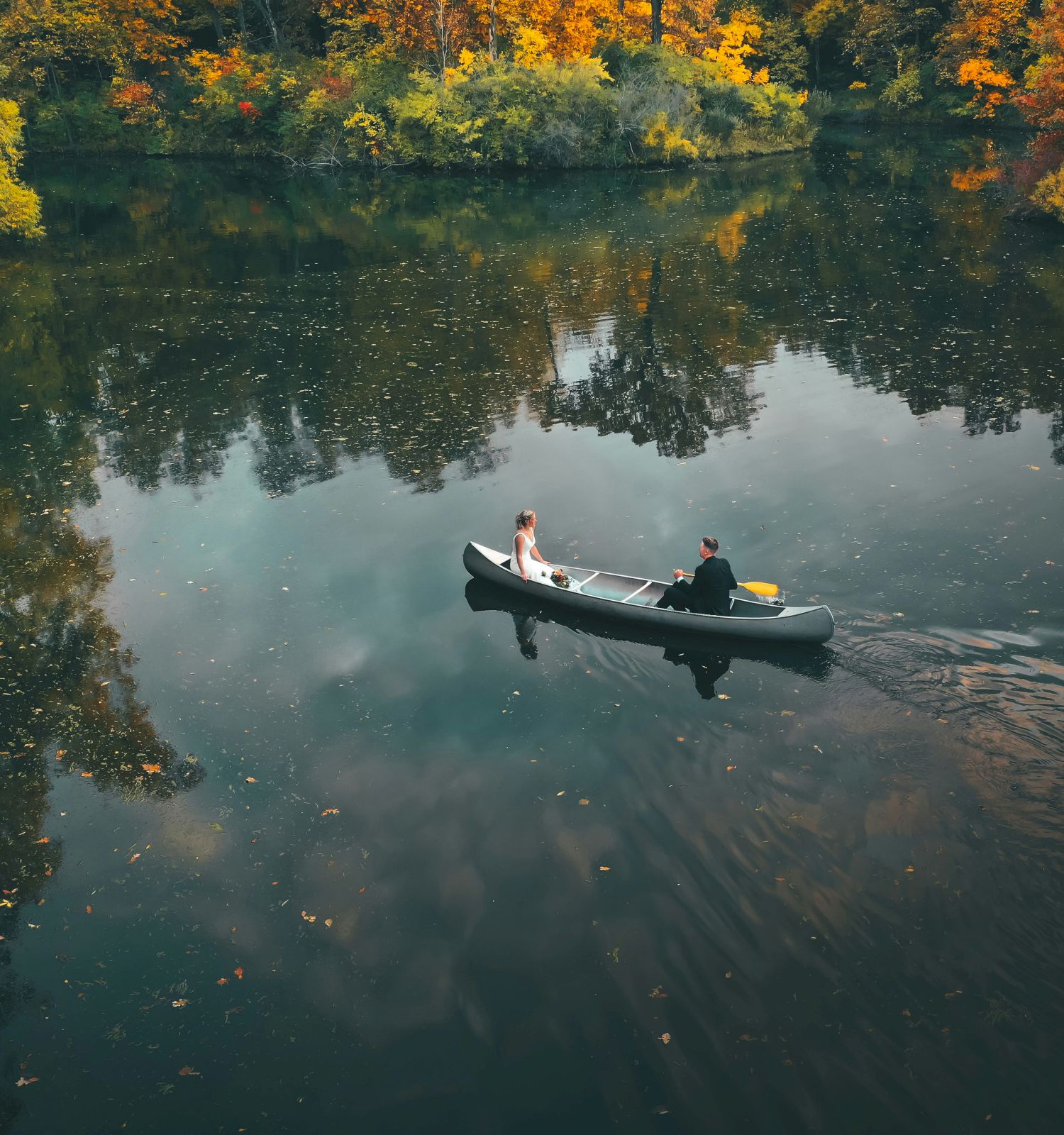 Two people are rowing a boat on a calm lake surrounded by trees with autumn foliage, reflecting on the water.