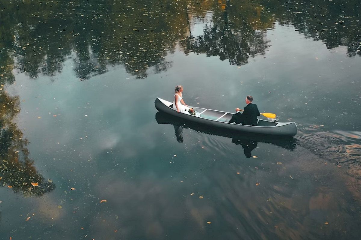 Two people in formal attire are in a canoe on a calm body of water with reflections of trees and sky, creating a serene, picturesque scene.