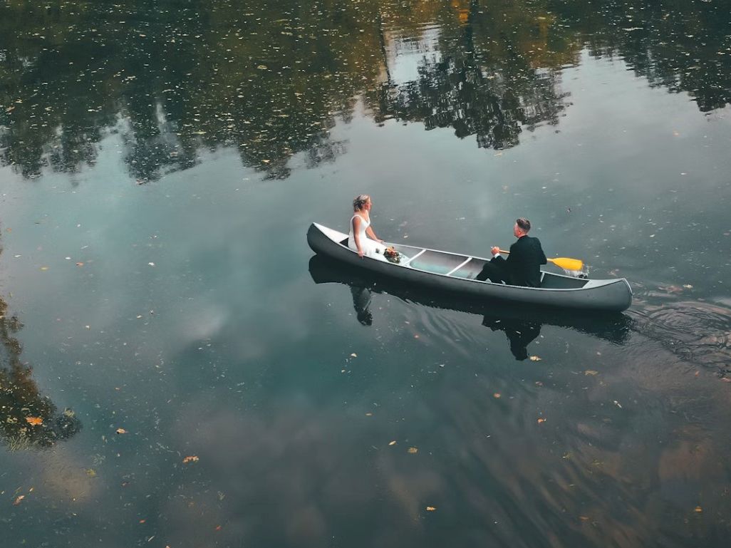 Two people in formal attire are in a canoe on a calm body of water with reflections of trees and sky, creating a serene, picturesque scene.