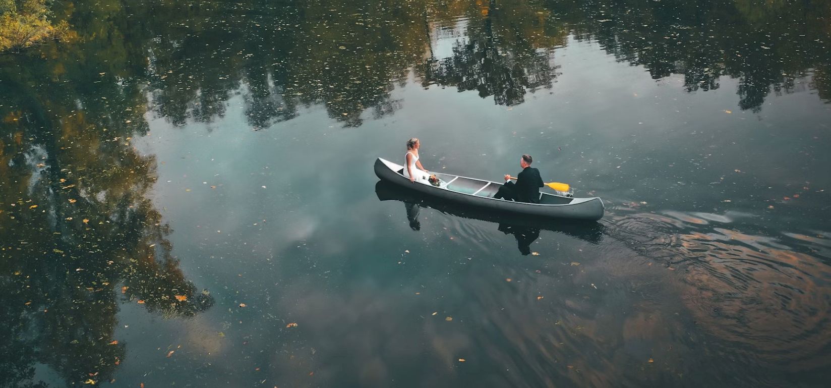 Two people in formal attire are in a canoe on a calm body of water with reflections of trees and sky, creating a serene, picturesque scene.